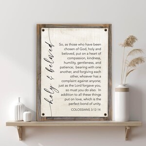 May include: A rustic wooden sign with a white background and black text. The sign features a Bible verse from Colossians 3:12-14 about being holy and beloved. The text reads: "So, as those who have been chosen of God, holy and beloved, put on a heart of compassion, kindness, humility, gentleness, and patience; bearing with one another, and forgiving each other, whoever has a complaint against anyone; just as the Lord forgave you, so must you do also. In addition to all these things put on love, which is the perfect bond of unity. COLOSSIANS 3:12-14"