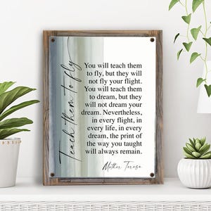 May include: A framed print with a wooden border features a quote by Mother Teresa. The text, in black script, is set against a watercolor background of blue, green, and white. The words "Teach them to fly" are written vertically on the left.