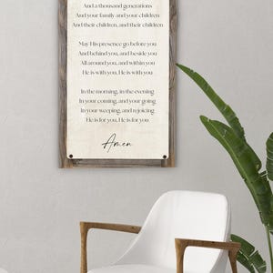 May include: A framed wooden sign with the text "The Blessing" in elegant script. The sign features a prayer, with the word "Amen" at the bottom. A white chair with wooden legs and a plant in a white pot are in the foreground.