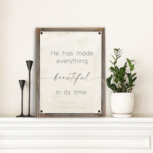 May include: A white wooden sign with a black inscription that reads "He has made everything beautiful in its time. Something Promised". The sign is hanging on a white wall above a white mantelpiece.