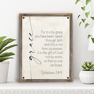 May include: A white wooden sign with a black distressed wood frame. The sign has a black and white quote about grace from Ephesians 2:8-9. The quote reads: "For it is by grace you have been saved. through faith -and this is not from yourselves. it is the gift of God -not by works. so that no one can boast. Ephesians 2:8-9"