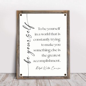 May include: A framed wall art piece with a rustic wooden frame. The artwork features the words "be yourself" in cursive script, with a quote by Ralph Waldo Emerson in a classic font. The background is off-white.