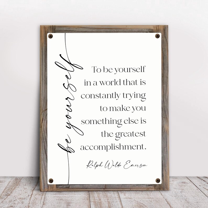 May include: A framed wall art piece with a rustic wooden frame. The artwork features the words "be yourself" in cursive script, with a quote by Ralph Waldo Emerson in a classic font. The background is off-white.