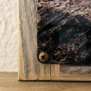 May include: Close-up of a wooden frame with a screw securing a picture of a rocky landscape. The frame has a rustic, weathered look.