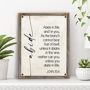 May include: A rustic wooden sign with a white background and black text. The text reads "Abide in Me, and I in you. As the branch cannot bear fruit of itself, unless it abides in the vine, neither can you, unless you abide in Me. JOHN 15:4"