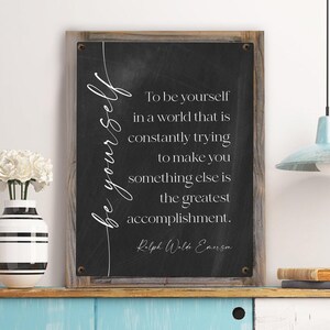 May include: A framed chalkboard sign with the quote "To be yourself in a world that is constantly trying to make you something else is the greatest accomplishment." The sign has a wooden frame and is displayed on a shelf with a vase of white flowers.