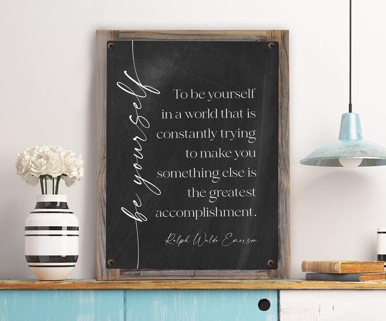 May include: A framed chalkboard sign with the quote "To be yourself in a world that is constantly trying to make you something else is the greatest accomplishment." The sign has a wooden frame and is displayed on a shelf with a vase of white flowers.