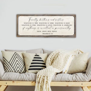 May include: A rectangular wooden sign with a light-colored background and black text. The text reads "Finally, brothers and sisters... Think about such things. Philippians 4:8." The sign is mounted above a sofa with decorative pillows and a cream-colored throw.