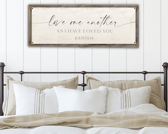 May include: A wooden sign with a white background and a black inscription that reads "love one another as I have loved you JOHN 13:34". The sign is hanging above a bed with white and beige bedding.
