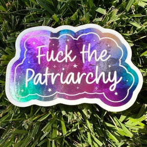 May include: A holographic sticker with a cloud-like shape and the text "Fuck the Patriarchy" in white script. The sticker has a colorful background with shades of purple, blue, and green, and small star accents.