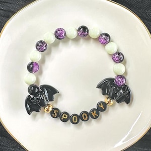 May include: A beaded bracelet featuring alternating purple crackle beads and pale green beads. The bracelet includes two black bat-shaped charms and black circular beads with gold lettering that spells out the word SPOOKY, accented by gold beads.