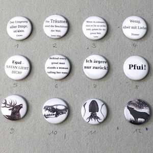Op de afbeelding: Twaalf ronde witte knoppen met zwarte tekst en afbeeldingen. De knoppen tonen verschillende citaten, spreuken en afbeeldingen, waaronder een hert, een schedel, een inktvis en een wolf.