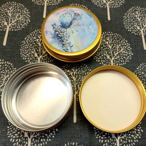 Könnte beinhalten: Zwei leere Metallbehälter mit goldenen Deckeln. Ein Deckel hat ein Aquarelldesign eines blauen Schmetterlings auf einem Lavendelhintergrund. Der andere Deckel ist schlicht.