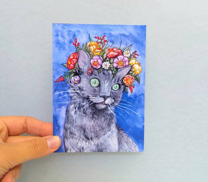 May include: A watercolor painting of a gray cat wearing a colorful flower crown. The cat has green eyes and a sweet expression. The background is a light blue watercolor wash.