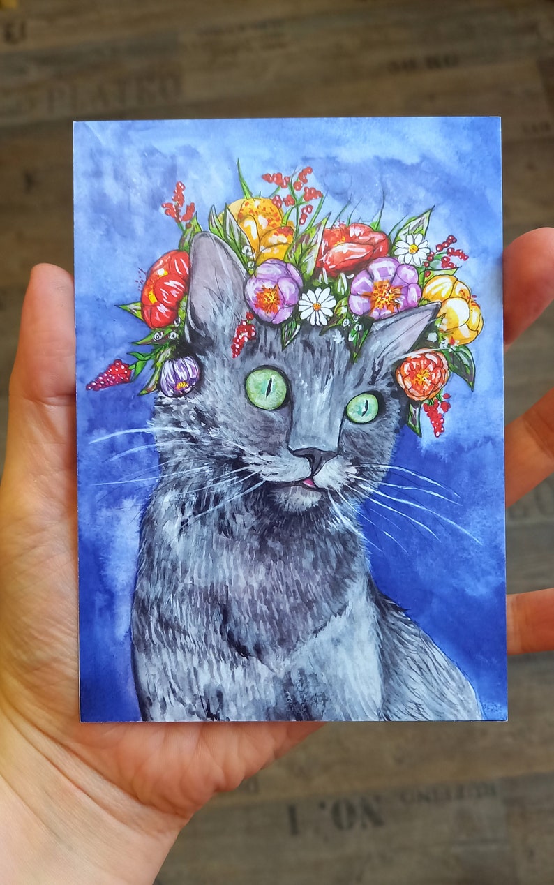 May include: A gray cat with green eyes wearing a flower crown. The crown is made of various flowers in shades of red, yellow, pink, and white. The cat is looking directly at the viewer with a slightly surprised expression. The background is a watercolor wash of blue.