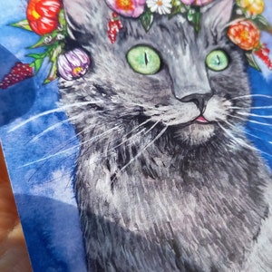 May include: A watercolor painting of a gray cat wearing a flower crown. The cat has green eyes and a pink tongue sticking out. The background is a blue watercolor wash.