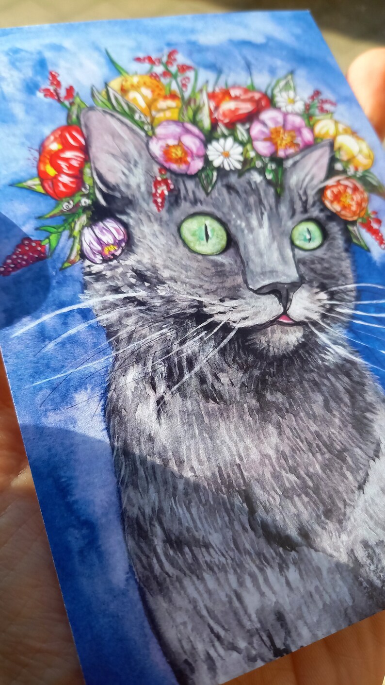 May include: A watercolor painting of a gray cat wearing a flower crown. The cat has green eyes and a pink tongue sticking out. The background is a blue watercolor wash.