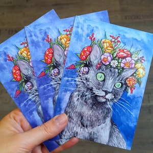 May include: Three watercolor paintings of a gray cat wearing a flower crown. The cat has green eyes and a pink nose. The flowers are red, yellow, orange, and purple. The background is a light blue watercolor wash.