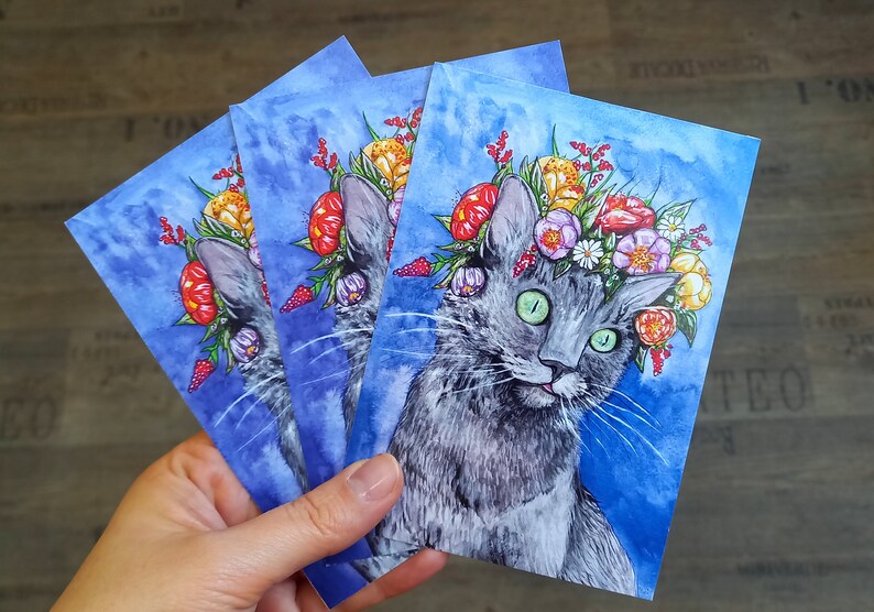 May include: Three watercolor paintings of a gray cat wearing a flower crown. The cat has green eyes and a pink nose. The flowers are red, yellow, orange, and purple. The background is a light blue watercolor wash.