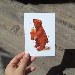 Pode incluir: Uma ilustração de desenho animado de um urso marrom segurando um favo de mel amarelo e laranja. O urso está olhando para cima e há pequenas abelhas pretas voando ao redor dele. O texto "Bär" está escrito abaixo do urso.