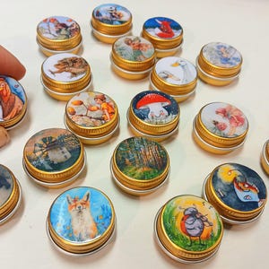 May include: A collection of small, round, gold-colored tins with various illustrations on their lids. The lids feature images of animals, landscapes, and whimsical scenes. One tin is held by a hand. The tins are arranged on a white surface.