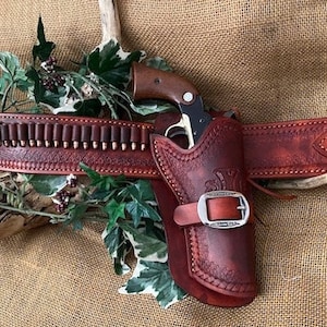 May include: A brown leather gun belt with a holster and a silver buckle. The belt has a cartridge loop and is decorated with stitching. The holster is designed to hold a small revolver.