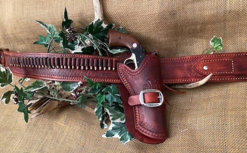 May include: A brown leather gun belt with a holster and a silver buckle. The belt has a cartridge loop and is decorated with stitching. The holster is designed to hold a small revolver.