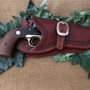 May include: A brown leather holster with a silver buckle and a black and silver revolver inside. The holster has decorative stitching and is lying on a hessian surface with green leaves.