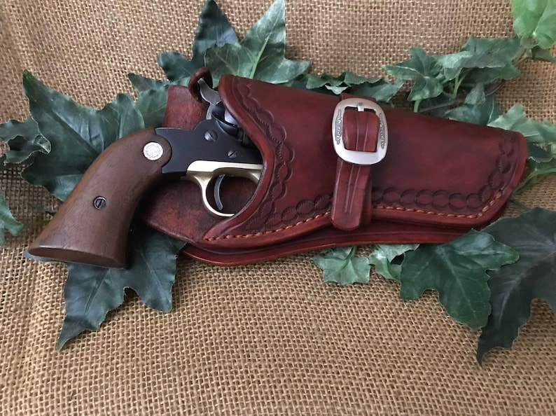 May include: A brown leather holster with a silver buckle and a black and silver revolver inside. The holster has decorative stitching and is lying on a hessian surface with green leaves.