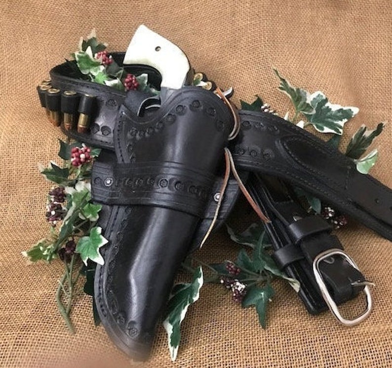 May include: A black leather holster with intricate stitching and a white-handled revolver. The holster is attached to a black leather belt with a silver buckle.