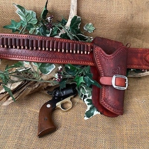 May include: A brown leather gun belt with a holster and a cartridge belt. The belt has a silver buckle and is decorated with stitching. A small revolver is lying on the belt.