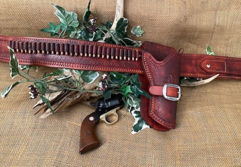 May include: A brown leather gun belt with a holster and a cartridge belt. The belt has a silver buckle and is decorated with stitching. A small revolver is lying on the belt.