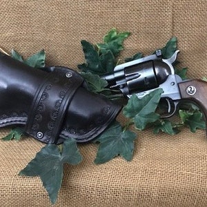 May include: A black leather holster with intricate stitching and a silver revolver with a wooden handle. The gun is in a holster and surrounded by green leaves.
