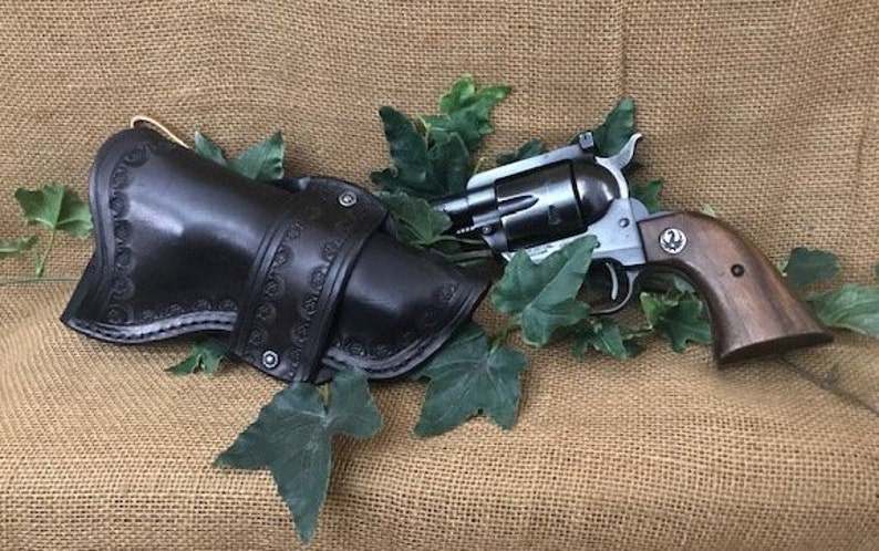May include: A black leather holster with intricate stitching and a silver revolver with a wooden handle. The gun is in a holster and surrounded by green leaves.