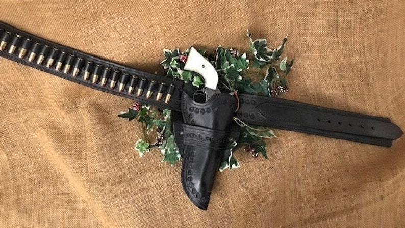 May include: A black leather gun belt with a holster and a silver and ivory revolver. The belt has a row of bullets attached to it.