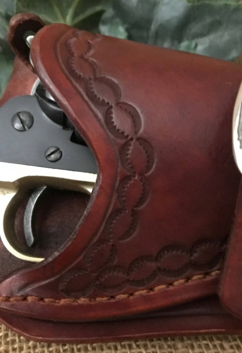 May include: A brown leather holster with a decorative pattern. A black and silver revolver is partially visible inside the holster.