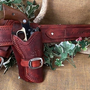 May include: A brown leather gun belt with a holster and a silver buckle. The holster has a brown leather flap with a silver buckle and the initials "S.Y." embossed on it. A silver revolver is in the holster.
