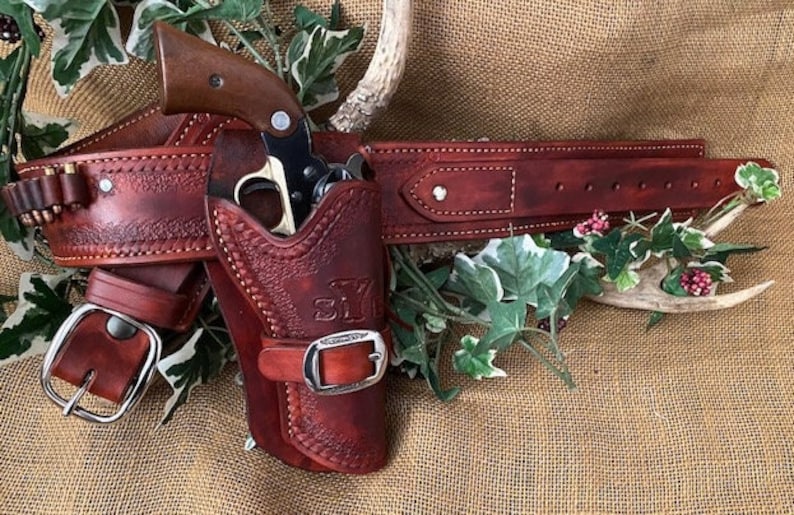 May include: A brown leather gun belt with a holster and a silver buckle. The holster has a brown leather flap with a silver buckle and the initials "S.Y." embossed on it. A silver revolver is in the holster.