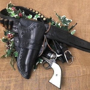 May include: A black leather gun holster with a silver buckle and a revolver with a pearl handle. The holster is decorated with stitching and a pattern of small circles. The revolver is lying on top of the holster.