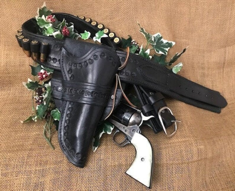 May include: A black leather gun holster with a silver buckle and a revolver with a pearl handle. The holster is decorated with stitching and a pattern of small circles. The revolver is lying on top of the holster.