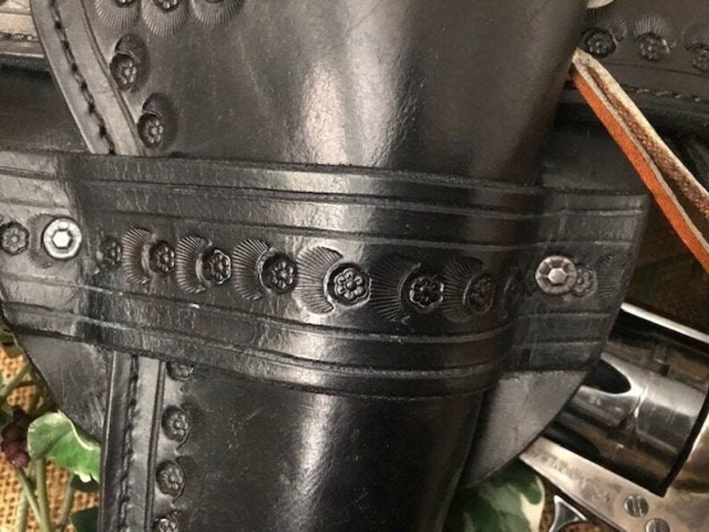 May include: A black leather holster with intricate floral designs and metal rivets. The holster is designed to hold a revolver.
