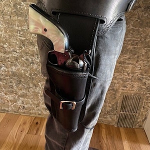 Puede incluir: Una funda de cuero negra con un revólver plateado y empuñadura de nácar. La funda se lleva en la cadera de una persona y se asegura con un cinturón.