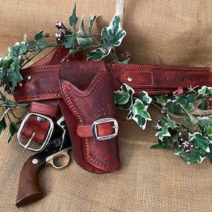 May include: A brown leather gun holster with a silver buckle and a matching belt. The holster is decorated with stitching and a floral design. The belt has a silver buckle and is also decorated with stitching.