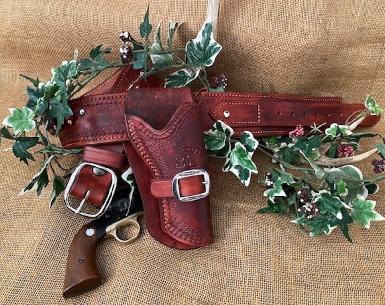 May include: A brown leather gun holster with a silver buckle and a matching belt. The holster is decorated with stitching and a floral design. The belt has a silver buckle and is also decorated with stitching.