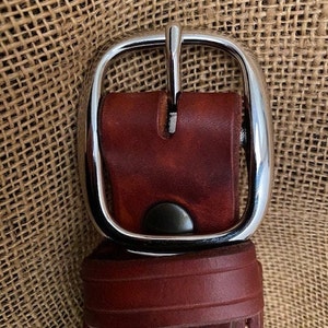 May include: A close-up of a brown leather belt with a silver buckle. The buckle is a simple, rounded design with a smooth finish.