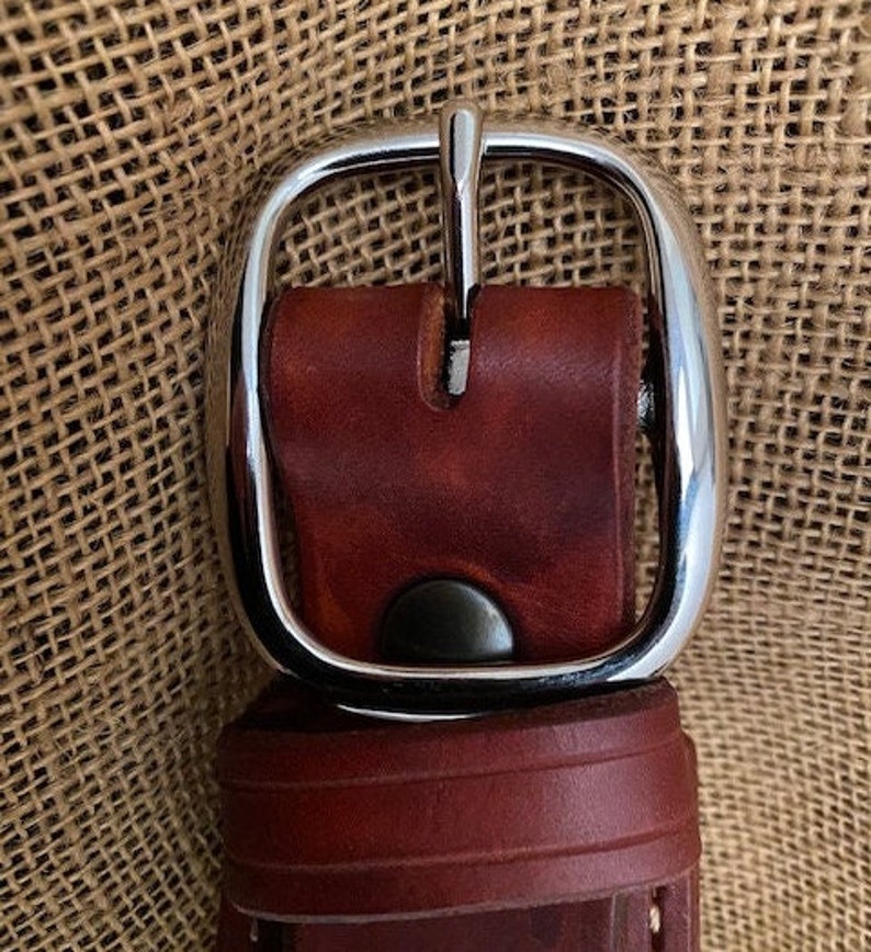 May include: A close-up of a brown leather belt with a silver buckle. The buckle is a simple, rounded design with a smooth finish.