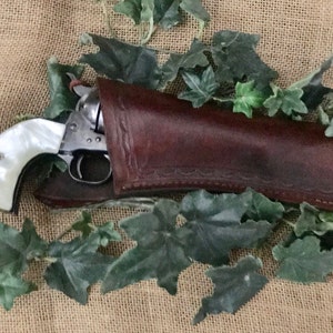 Può includere: Un revolver vintage con manico in perla è parzialmente inserito in una fondina di cuoio marrone. La pistola è argentata con un manico bianco. La fondina è marrone e presenta cuciture decorative. Foglie verdi circondano la pistola e la fondina.