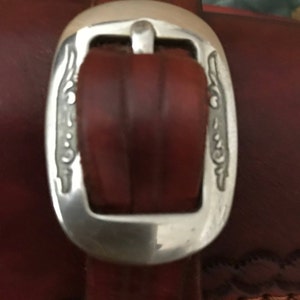 May include: Close-up of a brown leather belt with a silver buckle. The buckle has a decorative design on both sides.