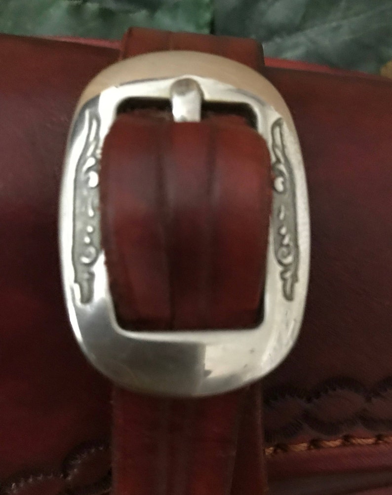 May include: Close-up of a brown leather belt with a silver buckle. The buckle has a decorative design on both sides.