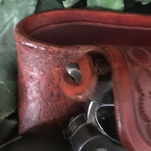 May include: Close-up of a brown leather holster with a metal snap closure. The holster is designed to hold a firearm.